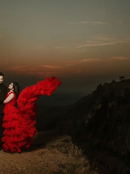 A wide-angle shot capturing the grandeur of a mountain pre-wedding location. The bride's ruffled red dress adds a pop of color to the dusky, atmospheric landscape.