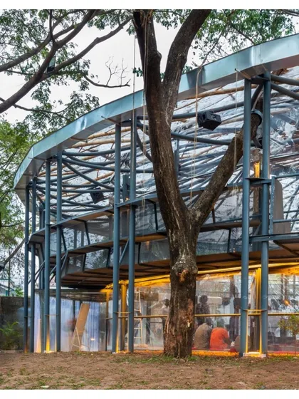 A view of the Koodaaram Pavilion's exterior, showing how the semi-transparent structure integrates with the surrounding trees at Cabral Yard in Kochi.