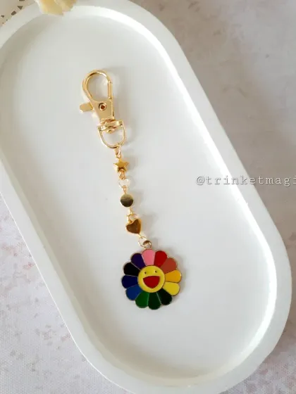 This bag charm features a colorful smiling flower, inspired by the artist Takashi Murakami. It's a bright and happy accessory to add to your collection.