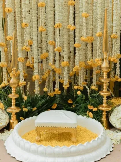 A detailed view of the main Haldi ceremony seat. The white stool is placed within a decorative base filled with marigold petals, surrounded by ornate brass lamps and floral arrangements.