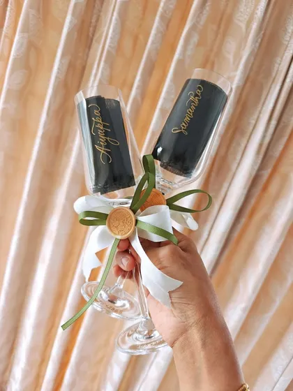 Holding up a pair of freshly engraved champagne flutes for 'Aiyappa' and 'Samantha'. The elegant script and custom ribbons make these a beautiful and personal wedding gift, ready to be part of their celebration.