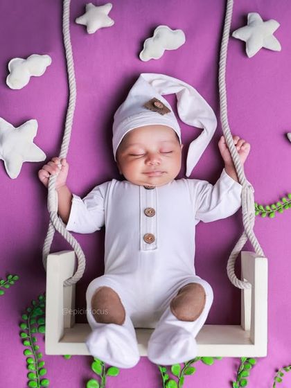 A whimsical setup with a baby in a sleeper cap on a swing against a vibrant purple backdrop, surrounded by stars, clouds, and greenery for a fantasy feel.