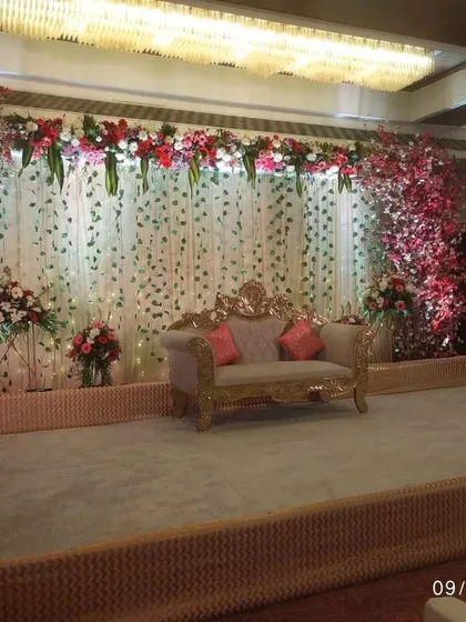 An elegant stage design for a wedding reception. The setup includes a beautiful backdrop of white floral strings, flanked by lush red and white flower arrangements and a classic, ornate sofa for the couple.