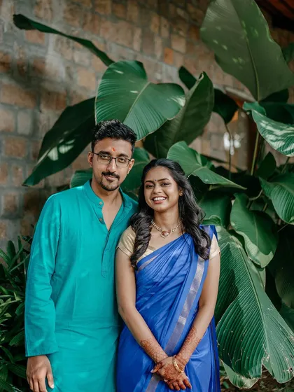 A lovely portrait of a couple in bright traditional South Indian attire, standing against a backdrop of large green leaves.