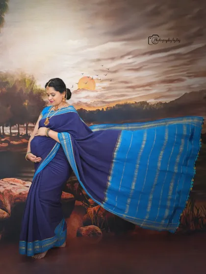 A beautiful studio portrait of a mother-to-be in a blue saree, with the fabric flowing elegantly against a painted backdrop.