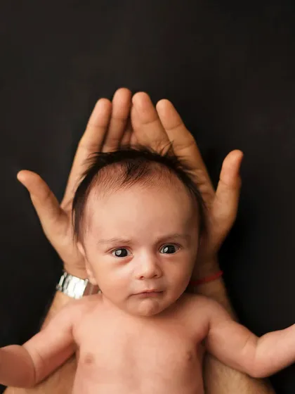 A newborn held in their parent's hands, looking directly at the camera. The dark background makes the baby the absolute center of attention.