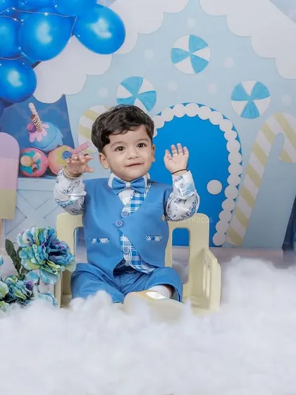 A happy wave from the birthday boy. This shot shows the full candy-themed set, complete with a balloon arch and fluffy clouds, designed for a magical first birthday photoshoot.