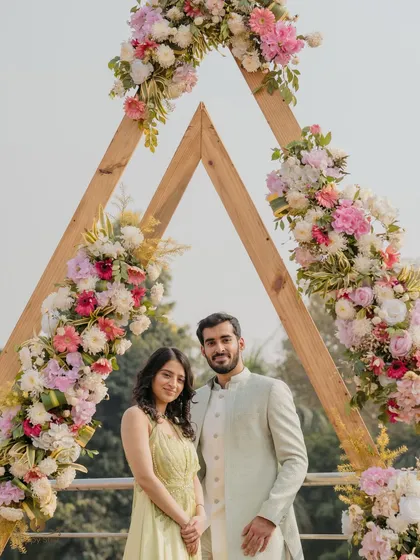 Karan and Satakshi posing under a beautiful triangular floral arch at their daytime Roka ceremony. The decor and their pastel outfits create a fresh and modern look.