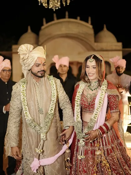 The happy couple walking hand-in-hand. Her royal red lehenga is perfectly complemented by our signature soft glam makeup.