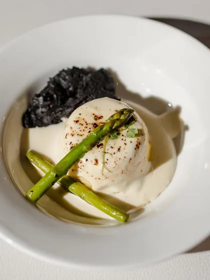 A dish of poached egg with asparagus and a black olive crumble. The composition is simple yet elegant, focusing on the quality of the ingredients and the balance of colors and textures.