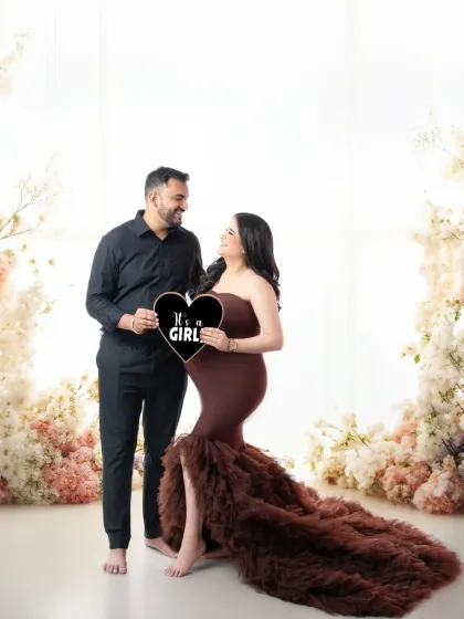 A joyful gender reveal photo for an expecting couple. They are surrounded by a beautiful floral setup as they announce "It's a Girl" with a heart-shaped sign.
