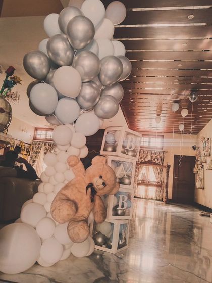 A cute welcome home decoration for a baby, featuring a large teddy bear climbing a column of white and silver balloons next to transparent "BABY" blocks.