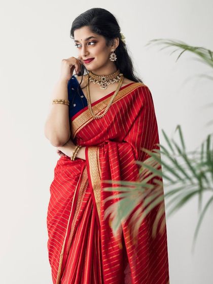 A three-quarter shot in a natural setting. The makeup looks fresh and vibrant, complementing the beautiful red and blue saree.