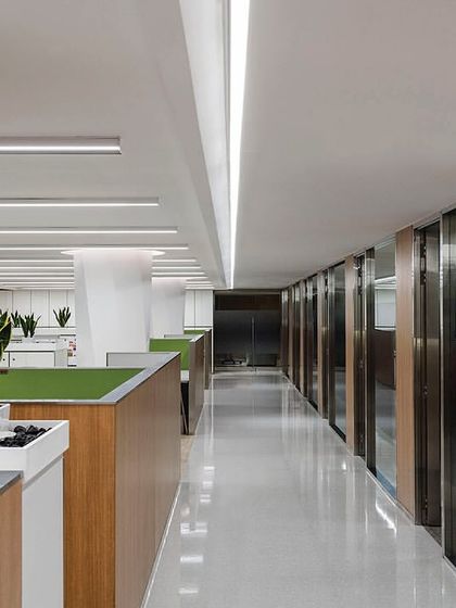A long corridor in 'The Layered Office' lined with private cabins on one side and open workstations on the other. The consistent use of wood paneling and integrated lighting creates a clean, unified aesthetic throughout the floor.