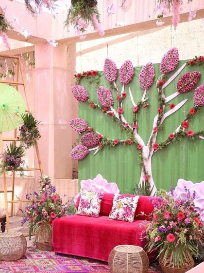 A creative and whimsical photo backdrop for a daytime event. A stylized tree made from floral arrangements stands against a pastel green wall, with a bright pink sofa and colorful floral baskets completing the look.