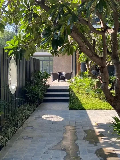 A tranquil pathway at a New Delhi farmhouse, shaded by a mature Plumeria tree. The path leads to a secluded seating area, demonstrating our approach to creating journey and discovery within a garden.