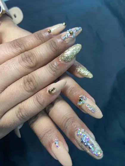 For when you want to make a statement. These long, almond-shaped nail extensions feature a mix of nude polish, dazzling gold glitter, and carefully placed rhinestones for a truly luxurious and festive feel.