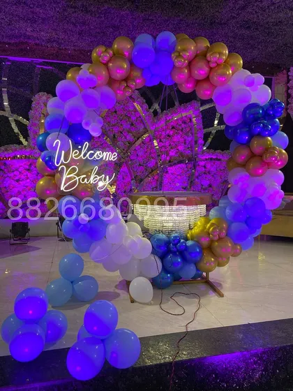 The welcome baby ring backdrop just after setup, showing the vibrant blue, white, and gold balloons and the bright glow of the neon sign.