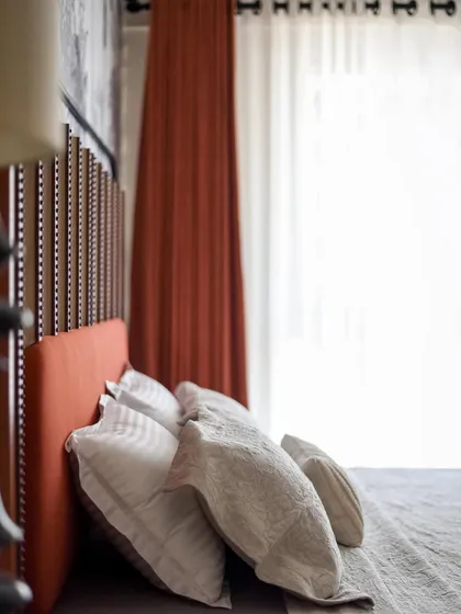 A side view of the bed, highlighting the texture of the headboard and the soft, inviting pillows. The warm orange curtains complement the color scheme perfectly.