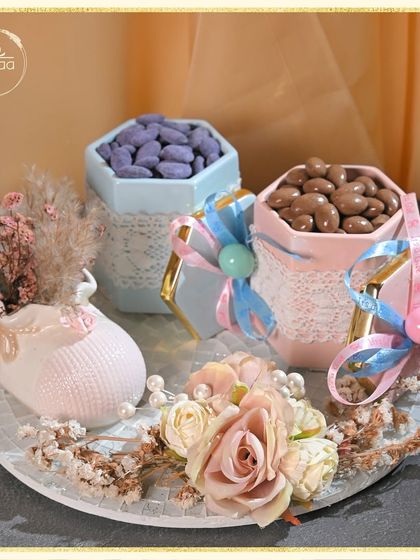 This gender reveal hamper is designed with both pink and blue elements. It features a ceramic baby shoe vase, dried flowers, and jars of pink and blue candies, making it a fun and stylish way to share the surprise.
