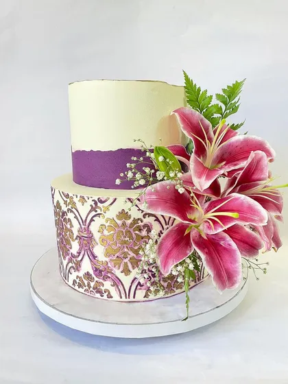 Artistry and flavor come together in this two-tier engagement cake. I used a custom stencil to create a detailed damask pattern in purple and gold on the bottom tier, contrasting it with a clean top tier and a vibrant arrangement of fresh stargazer lilies.