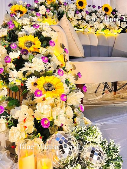 A close-up of the floral couch decor, showing the mix of sunflowers, daisies, and purple accent flowers. The addition of disco balls and candles at the base adds a fun and glamorous touch.