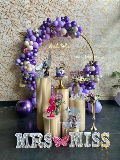 A sophisticated lavender-themed bachelorette party. The setup includes a golden ring arch with purple and silver balloons, a "Bride to be" neon sign, and "MRS" and "MISS" marquee letters.