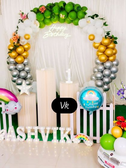 A soft and cheerful birthday setup with a circular balloon arch in green, gold, and silver. It includes a 'Happy Birthday' neon sign, a white picket fence, and light-up marquee letters.