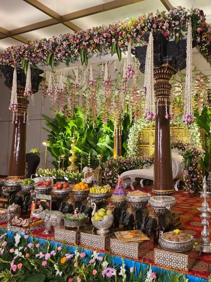 A closer view of the wooden pillar mandapam, highlighting the hanging jasmine garlands and the detailed offering table with cow and elephant props.