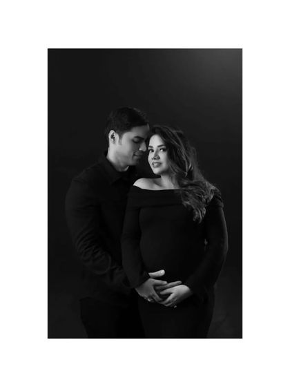 An intimate black and white maternity photo of a couple. His arms are wrapped around her, their hands resting on the baby bump, a classic portrait of love and anticipation against a dark background.