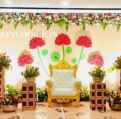A creative and modern stage for a half saree ceremony. The backdrop features stylized paper flowers and a royal throne, framed by traditional floral pillars.