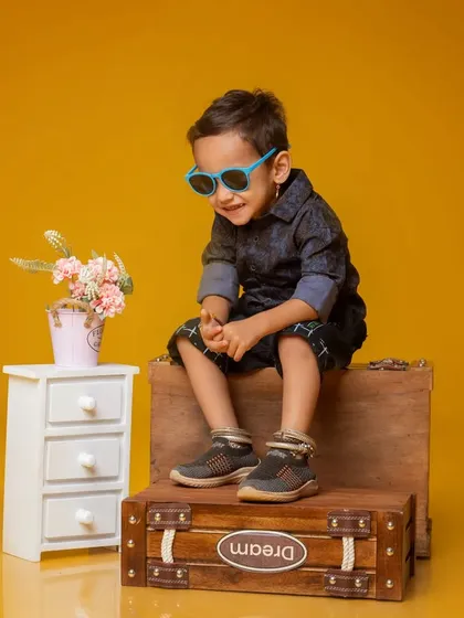 A candid moment of laughter from a third birthday session. The little boy, wearing sunglasses, enjoys the playful setup with a miniature dresser and 'Dream' suitcase.
