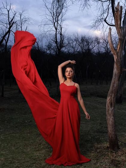 A dramatic and artistic pose. The flowing red fabric of the gown creates a stunning visual against the dark woods, perfect for a high-fashion pre-wedding shoot.