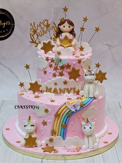 A dreamy two-tier pink cake for a birthday celebration. It's adorned with a fondant girl holding a star, cute unicorns, a rainbow slide, and golden star accents.