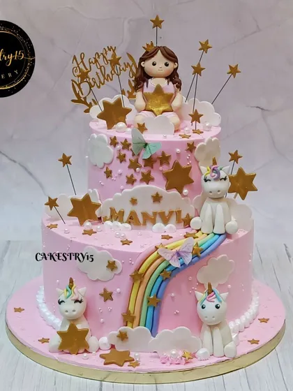 A dreamy two-tier pink cake for a birthday celebration. It's adorned with a fondant girl holding a star, cute unicorns, a rainbow slide, and golden star accents.