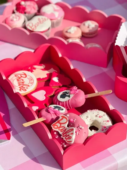 A tray of Barbie-themed sweets, including logo cookies, pink-drizzled donuts, and cakesicles, all presented in custom scalloped pink trays.