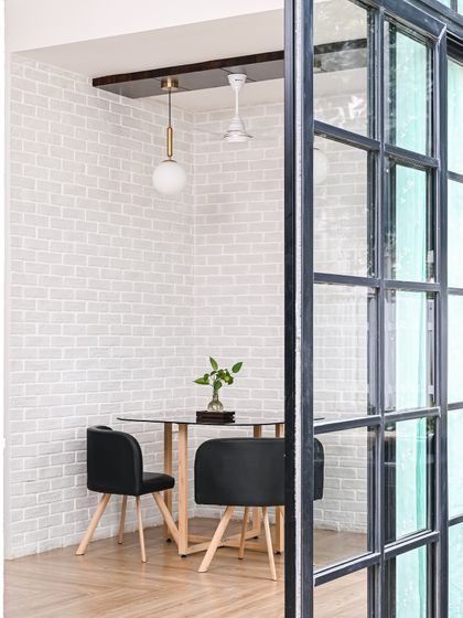 This intimate dining or meeting nook is defined by its textures. A white brick accent wall and herringbone wood flooring create a rustic-chic vibe, while the modern glass-top table and black chairs add a contemporary touch.