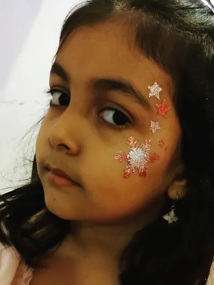This winter-themed face art features a large, sparkling snowflake surrounded by smaller stars. The use of white and pink glitter gives it a magical, frosty feel.