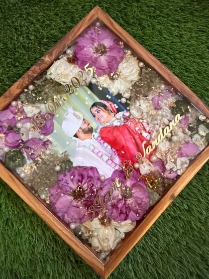 A diamond-oriented view of a varmala preservation frame. The wooden frame adds a warm, natural touch to the vibrant purple and white flowers preserved within.