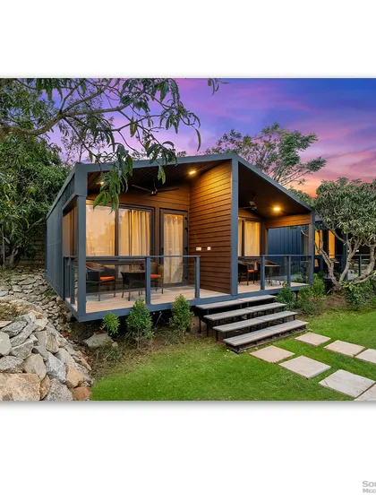 A pair of modern prefab cottages at dusk, nestled in a lush mango grove. The warm interior light and the dramatic sunset sky create a magical and inviting scene, showcasing the appeal of modular living.