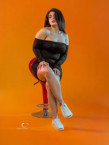 A classic, elegant pose on a stool. The combination of the little black dress, white sneakers, and vibrant background creates a chic and contemporary look.
