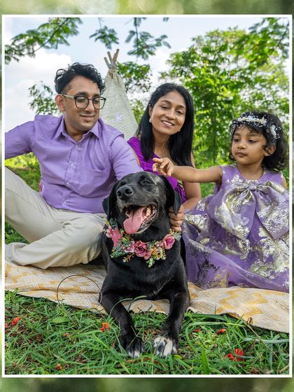 A happy family portrait in a lush green orchard. The family, dressed in shades of purple, smiles brightly with their black Lab, Bagheera, who is the clear star of the show with his happy pant and floral collar.