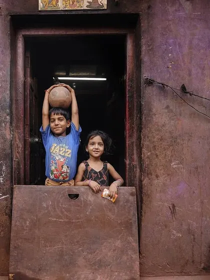 Two children peer out from their doorway, one playfully balancing a pot on his head. It's a candid, playful moment that offers a glimpse into family life within the community.