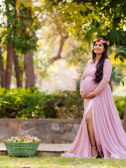 A joyful portrait of a mother-to-be in a pink gown and floral crown, standing next to a basket of flowers. The natural park setting and her happy expression create a fresh and vibrant image.