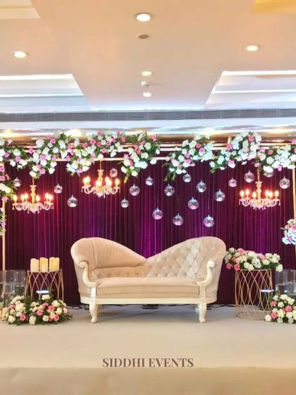 A sophisticated reception stage featuring a deep purple drape, hanging crystal baubles, and elegant chandeliers, all framed by a beautiful arch of white and pink florals.