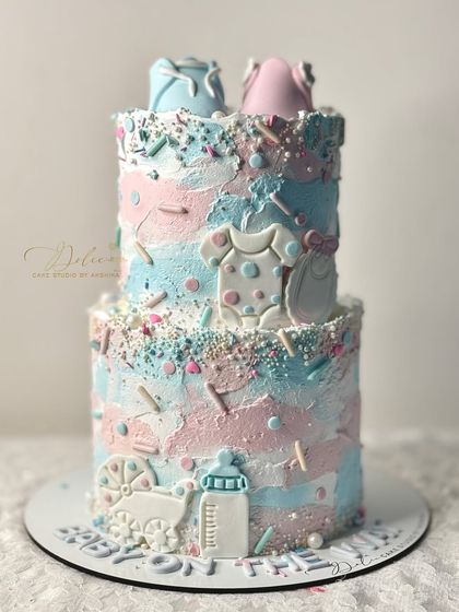 A "He or She" gender reveal cake with a textured pink and blue finish. It's decorated with cute fondant baby items like onesies, a bottle, and a carriage.