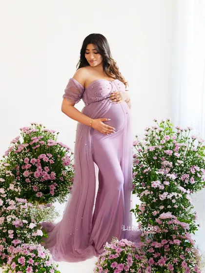 A full-length portrait of a mother-to-be surrounded by flowers. She stands gracefully in a lavender gown, her hands on her bump, amidst beautiful arrangements of purple flowers.
