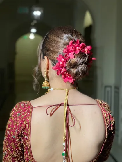 A textured low bun accented with vibrant pink orchids. This hairstyle adds a pop of color and a touch of the exotic, perfectly complementing the bride's red lehenga for her wedding ceremony.