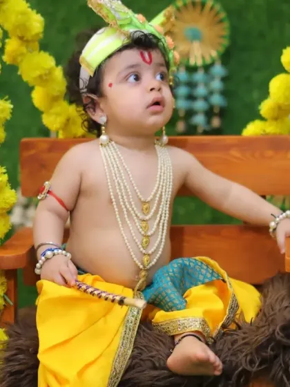 A beautiful, expressive portrait of a baby dressed as Krishna, seated on a fur-lined swing decorated with marigolds.