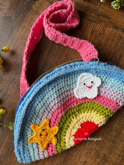 Our handmade rainbow crochet bag for kids is designed to bring a smile to any child's face. It features a colorful rainbow arc, cute star and cloud appliqués, and a bright pink strap.